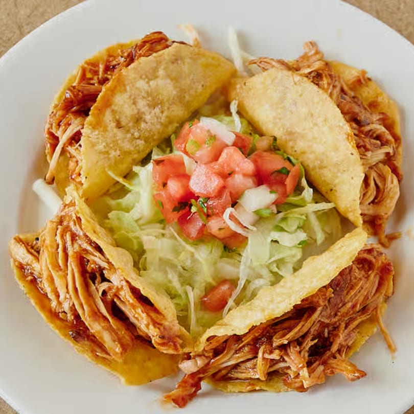 Four crispy tacos filled with shredded meat, surrounded by lettuce and pico de gallo.
