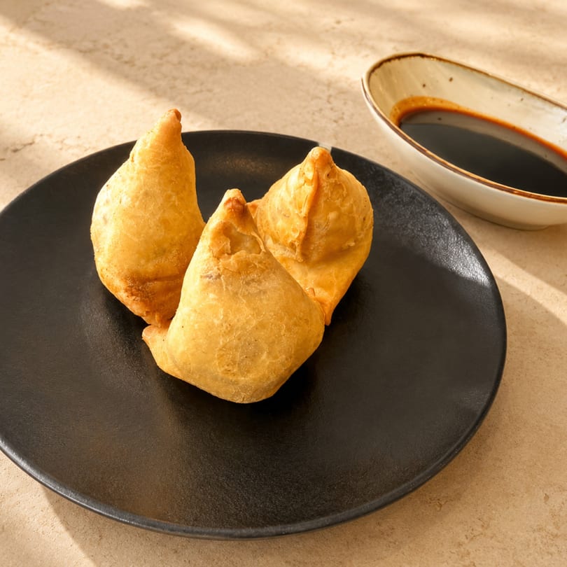 Three golden-brown samosas on a black plate, with a small dish of dark sauce beside them.