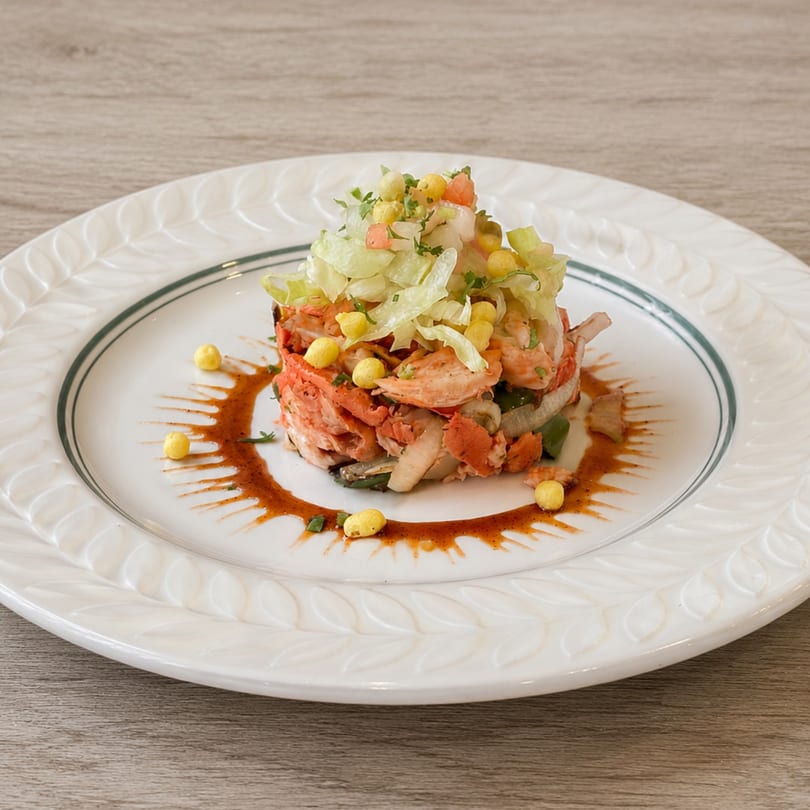 Lobster and shrimp ceviche tower topped with lettuce, corn, and pico de gallo on a white plate.